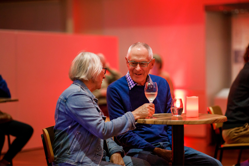 Two people seated at a small table during a social event, chatting over drinks in a warmly lit venue with red ambient lighting.