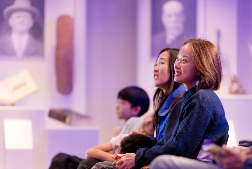 Audience members seated at a talk or presentation, watching attentively in a softly lit venue with exhibition displays in the background.