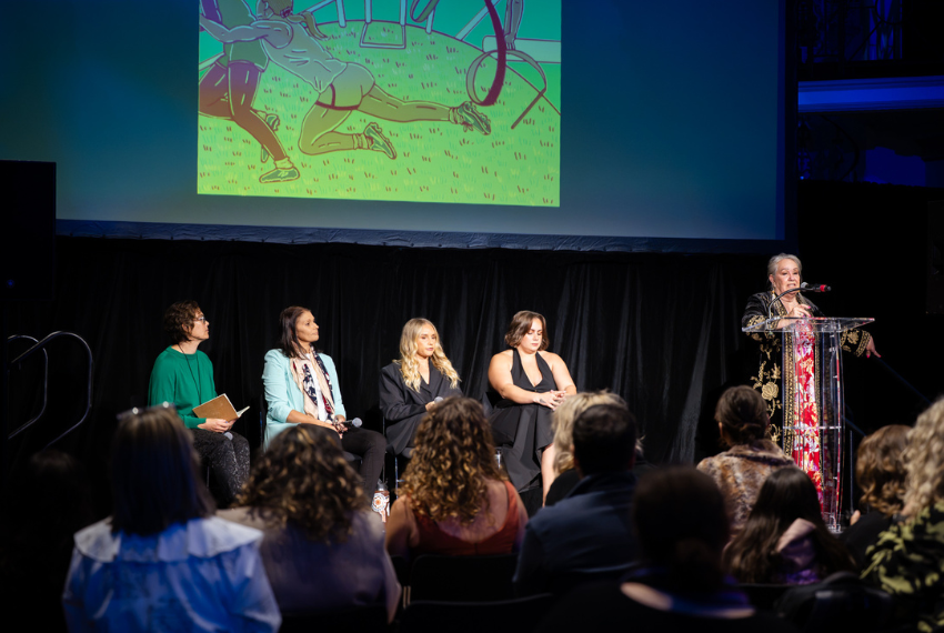 Panel discussion on a stage with four speakers seated and one presenter speaking at a lectern, while an audience listens and a large screen displays artwork above.