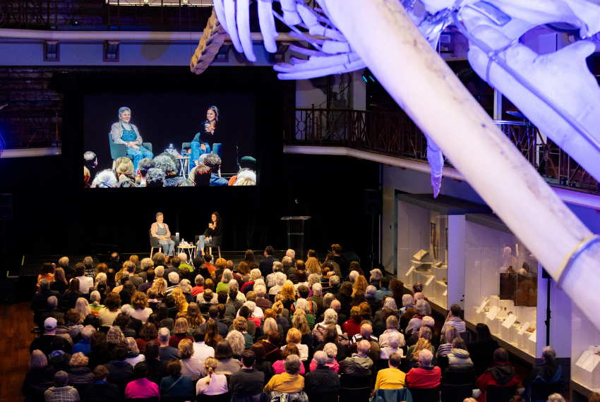 Large audience gathered for a live interview or discussion in a museum venue, with two speakers on stage and the conversation displayed on a large screen above.