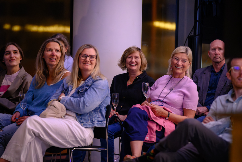 Audience members seated at an evening talk or event, listening to a speaker in a relaxed social setting with drinks in hand.