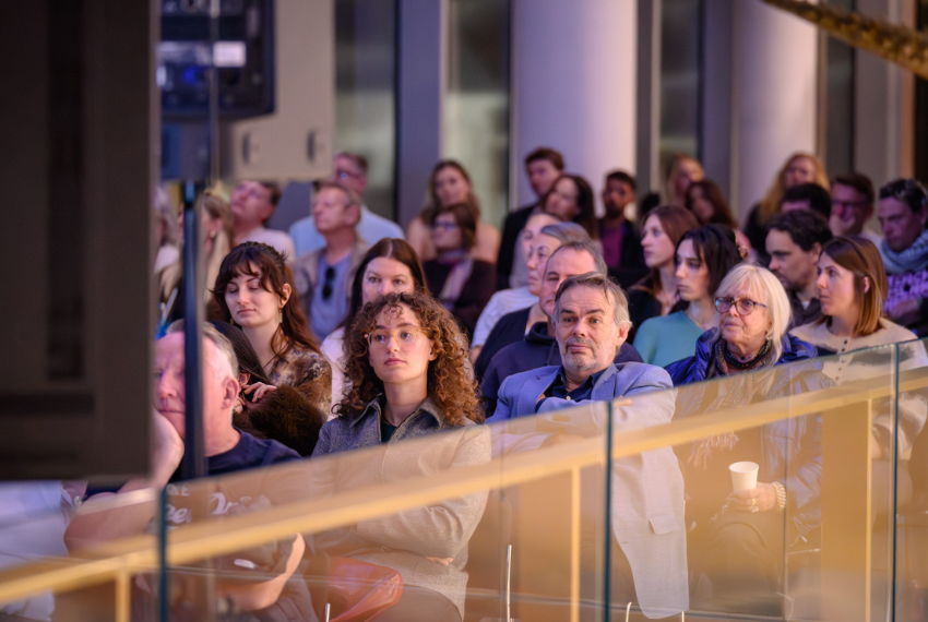 Audience members seated at a public talk in a museum or gallery space, listening attentively to a speaker off camera.
