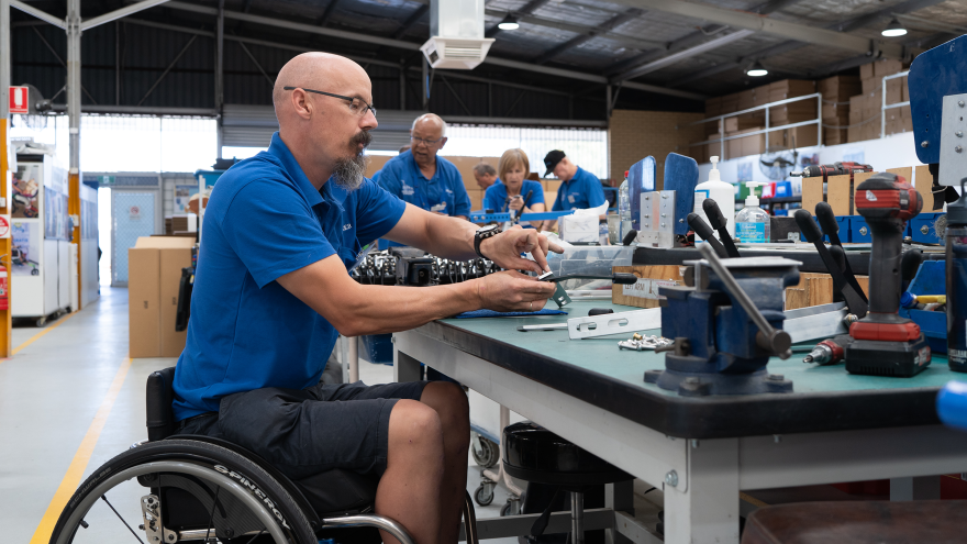 A wheelchair user assembles equipment at a workbench in a workshop, with tools, components and colleagues working in the background.