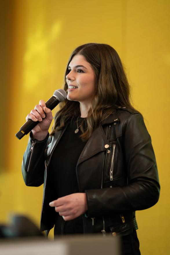 A woman with long brown hair, wearing a leather jacket, speaking into a microphone