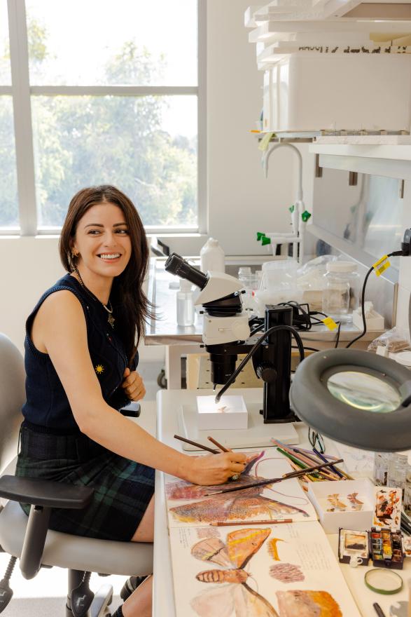 a person smiling who is sitting at a desk with a microscope with lots of instruments and tools around the desk including colourful illustrations
