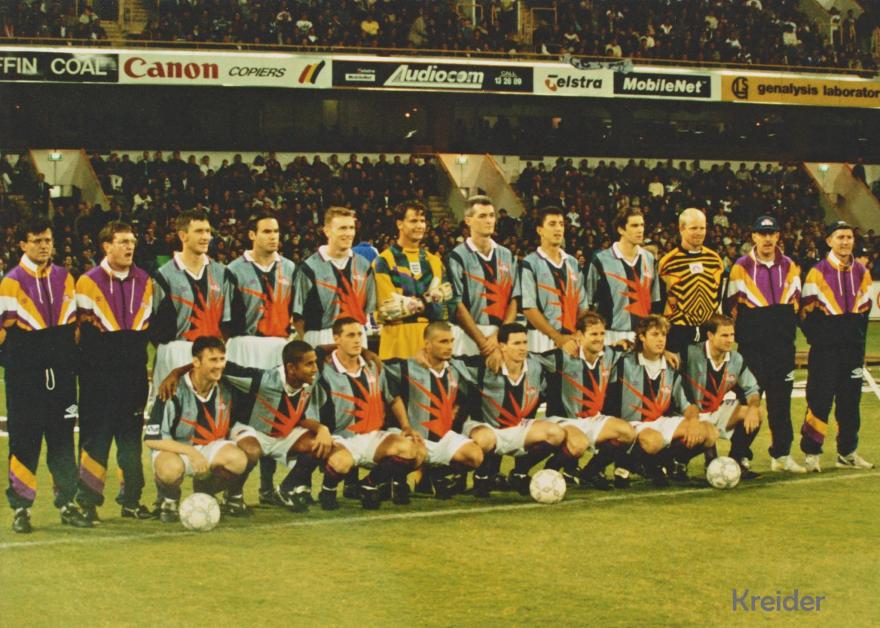 Perth Glory squad and coaching staff posing for a team photograph on the field before a match, with a crowded grandstand behind them