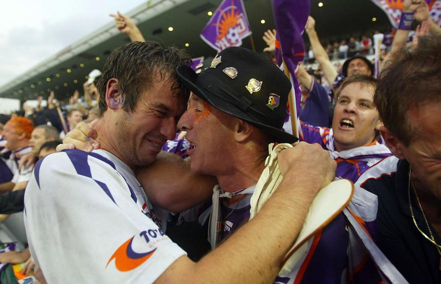 Perth Glory supporter embracing a player beside the pitch during post-match celebrations, with cheering fans and club flags in the background.