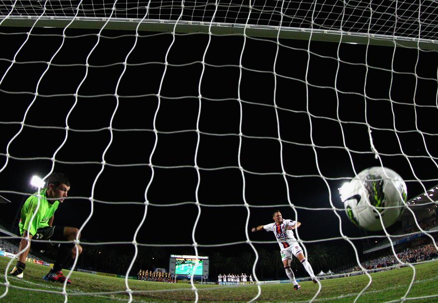 Footballer taking a penalty kick as the ball flies toward goal, viewed through the net from behind the goalkeeper