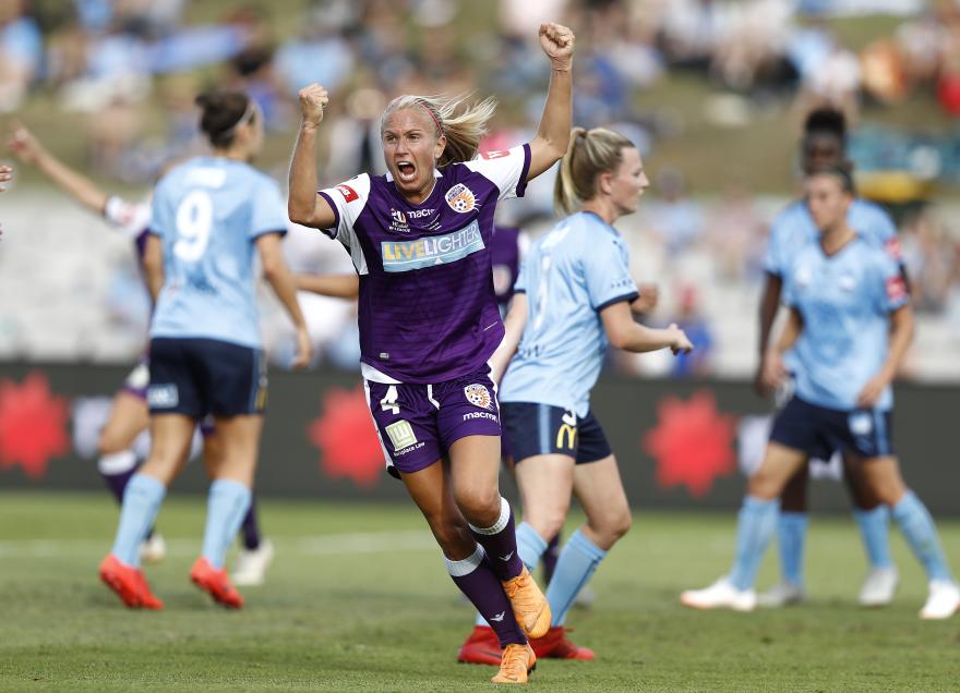 Player in a Perth Glory kit celebrates on the field with raised arms during a football match, with opposing players in the background.