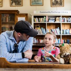 A child and adult wearing a cap sitting with a teddy bear at a desk, bookcases in the background