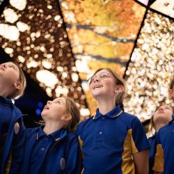 Four school children in blue uniforms stare up in awe at the colourful kaleidoscopic mineral tunnel at the WA Museum Boola Bardip