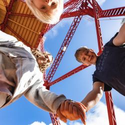 Two children hold hands and stand over the camera smiling down at the photographer while a third younger child peaks their head over into the show. Above them a large red metal structure looms large with a bright blue sky behind them