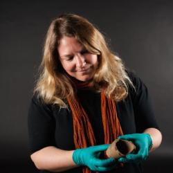 Annie Carson wears a black jumper and orange scarf with their blonde hair falling over their shoulders. They carefully hold a pottery fragment while wearing bright blue gloves and smiles down at it.