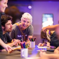 A small group of people sit at a table with paper and pencils in a colourful purple and blue lit gallery. A volunteer in a grey shirt leans over the table and smiles at the group
