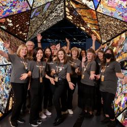 A small group of eleven people of various ages in matching black trousers and grey shirts with the word volunteer on the front, and gold lanyards. They all pose with their arms out, and stand under a large and colourful kaleidoscopic tunnel