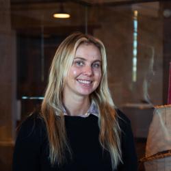 A person with long blonde hair smiles at the camera, wearing a dark jumper and collared shirt