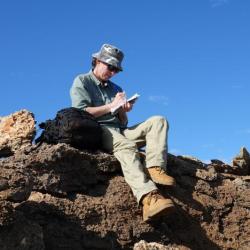 A person wearing a hat and long pants sits atop a rocky outcrop beneath a cloudless blue sky