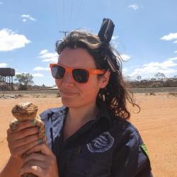 An image of a person wearing sunglasses and a dark jumper, holding a lizard in the Australian desert, beneath a blue sky