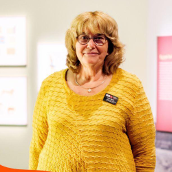 A person wearing a yellow top, with shoulder length hair and glasses, stands in a bright room.