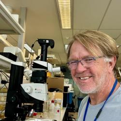 A person wearing glasses and a light blue shirt smiles at the camera, sitting beside a microscope