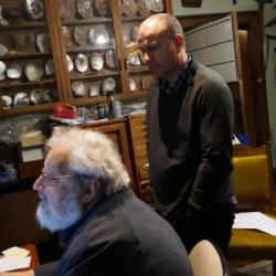 2 people, one sitting and one standing, looking in the same direction in a shadowy room, a cabinet of pearly shells in the background.