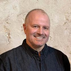 A person smiles at the camera wearing a dark collared shirt, standing in from of a limestone wall