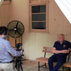 A person sits on a bench in front of a camera, which is being operated by a a person wearing a long sleeved, light blue shirt