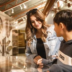 A young person in a grey hoodie and an adult with wavy brown hair and a denim jacket stand in a Museum gallery and stare down at a glass showcase with Aboriginal objects inside