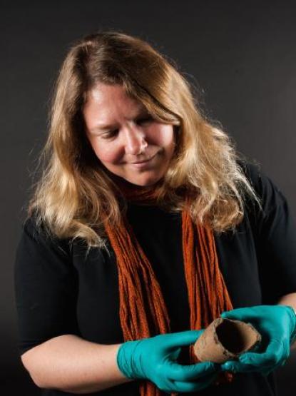 Annie Carson wears a black jumper and orange scarf with their blonde hair falling over their shoulders. They carefully hold a pottery fragment while wearing bright blue gloves and smiles down at it.