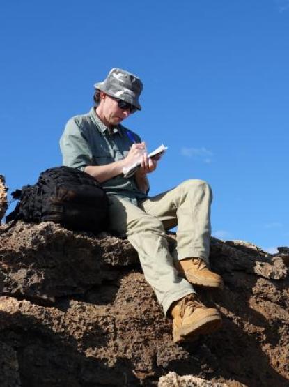 A person wearing a hat and long pants sits atop a rocky outcrop beneath a cloudless blue sky