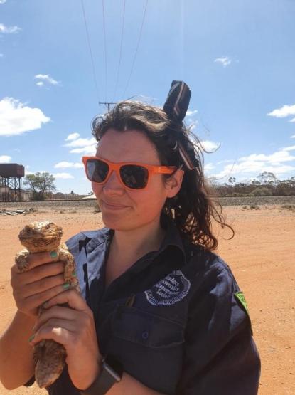 An image of a person wearing sunglasses and a dark jumper, holding a lizard in the Australian desert, beneath a blue sky