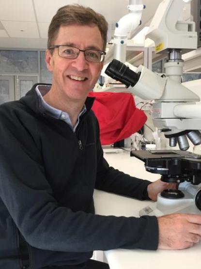 A person with short hair wearing glasses and a dark jumper sits in a science laboratory, next to a microscope.