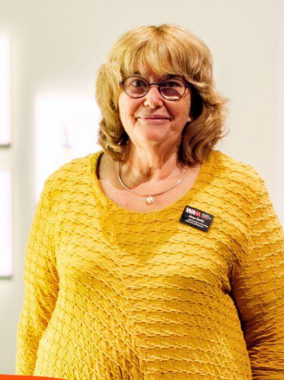 A person wearing a yellow top, with shoulder length hair and glasses, stands in a bright room.