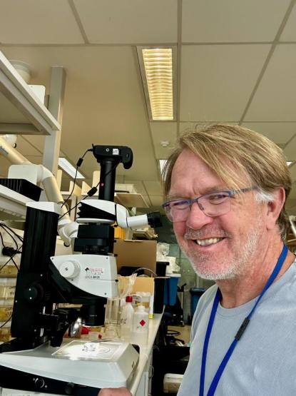 A person wearing glasses and a light blue shirt smiles at the camera, sitting beside a microscope