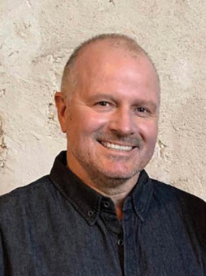 A person smiles at the camera wearing a dark collared shirt, standing in from of a limestone wall