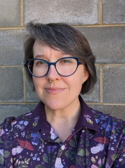 A person wearing blue glasses and a purple shirt with flowers and mushrooms stands in front of a brick wall