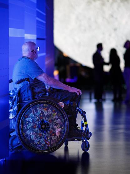 A person in a grey shirt and black glasses with a bald head sits in a wheel chair with an intricately decorated wheel featuring drawings of Australian birds and foliage. They are seated in a vividly-lit blue hallway with a futuristic design in front of a large moon sculpture