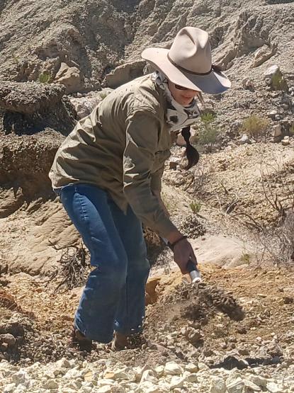 Person in a wide‑brim hat using a small trowel to dig in a rocky outdoor field site.