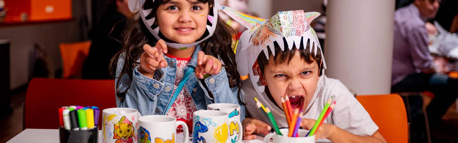 Image of two children roaring to the camera wearing paper dinosaur hats