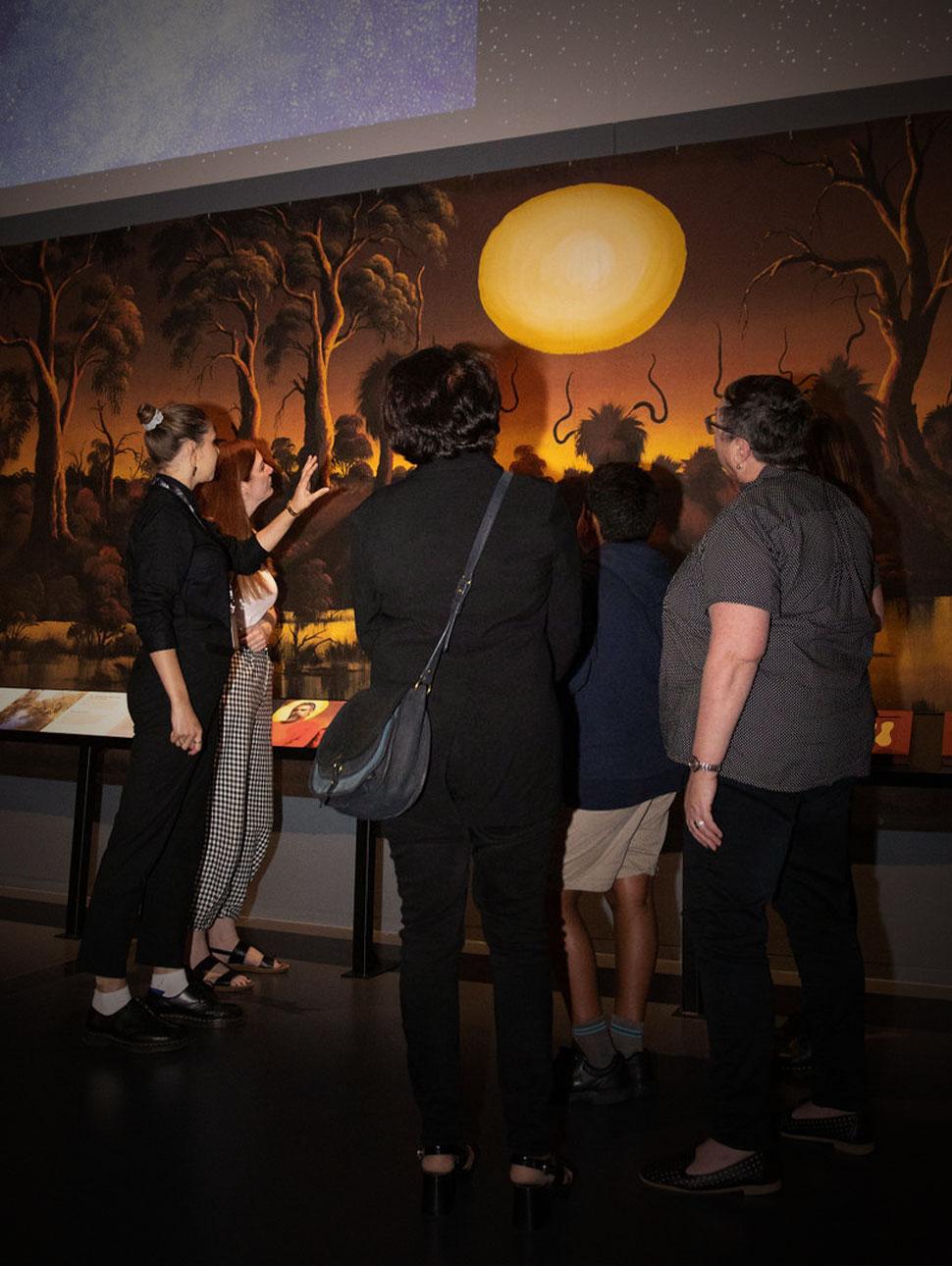 A group of people stand together with a tour guide, examining a large artwork in a museum gallery