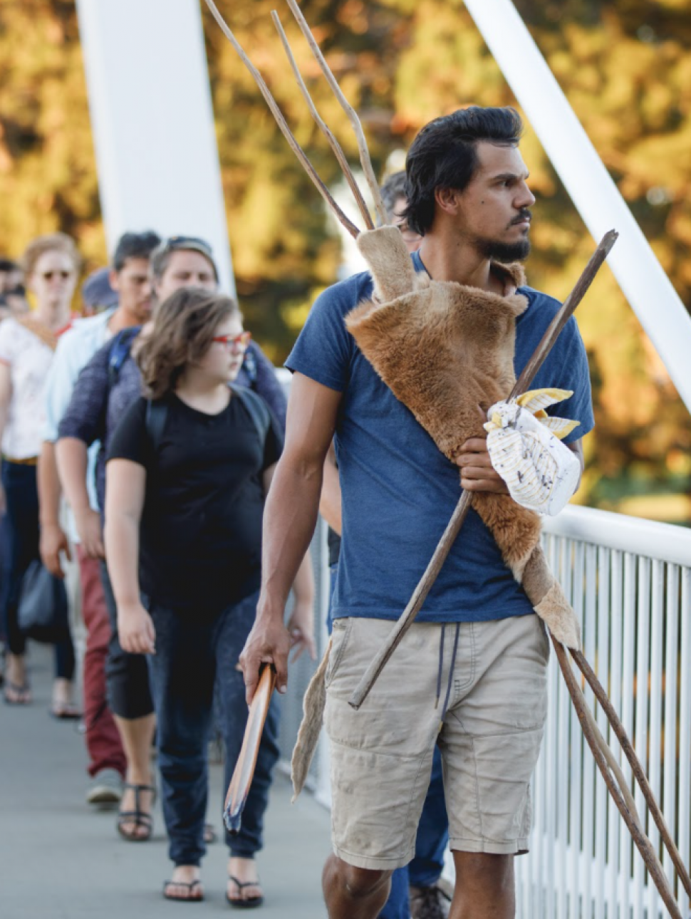 Several people are walking across a bridge towards the camera; the serious-looking man at the front is holding some wooden tools or weapons wrapped in an animal skin