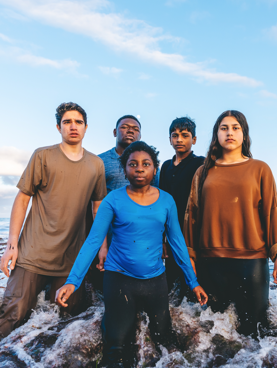 A diverse group of young people stand in the shallows of the ocean, looking seriously at the camera