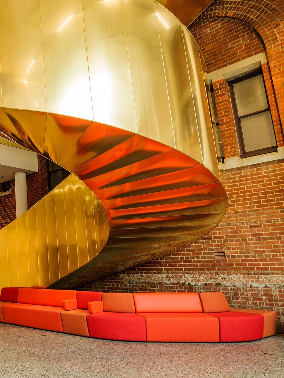 A golden spiral staircase winds upwards in front of a brick wall