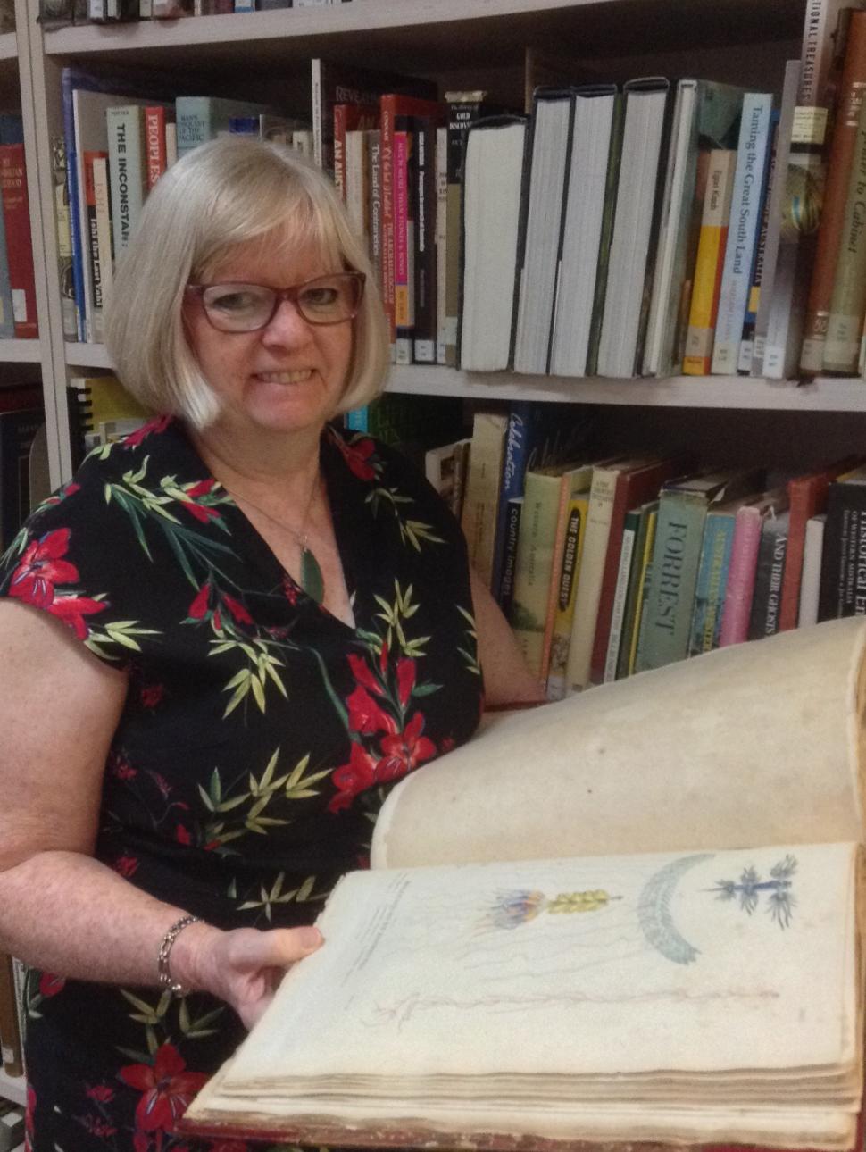 A woman stands in a library holding a large illustrated book