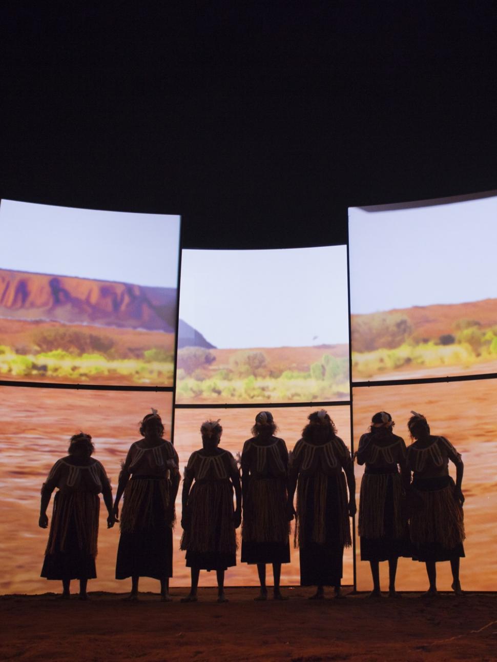 Artists from Songlines stand in front of a wall of screens showing a desert landscape