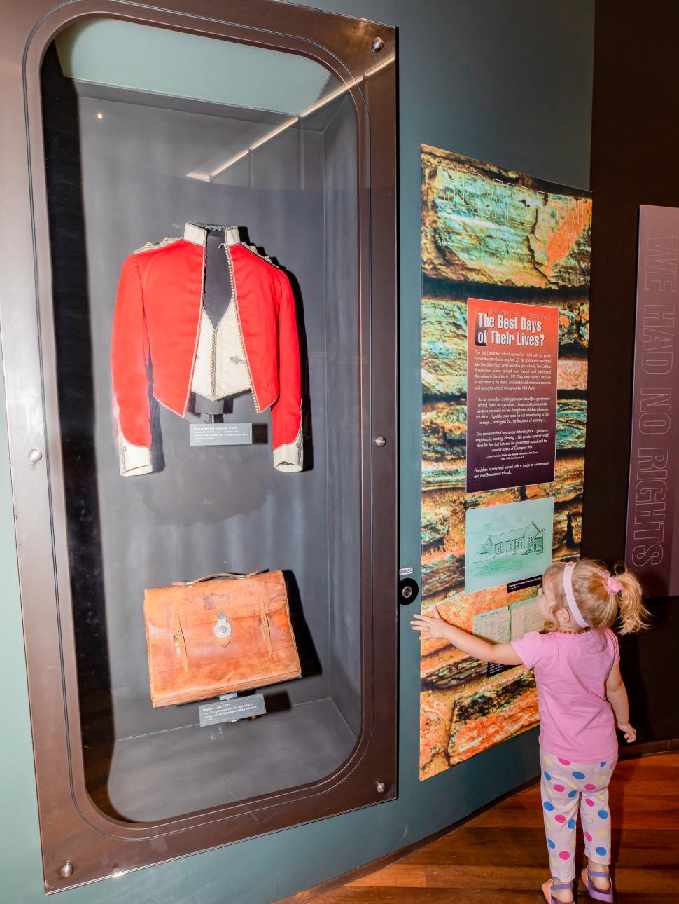A young girl examines a museum showcase containing an old jacket and bag