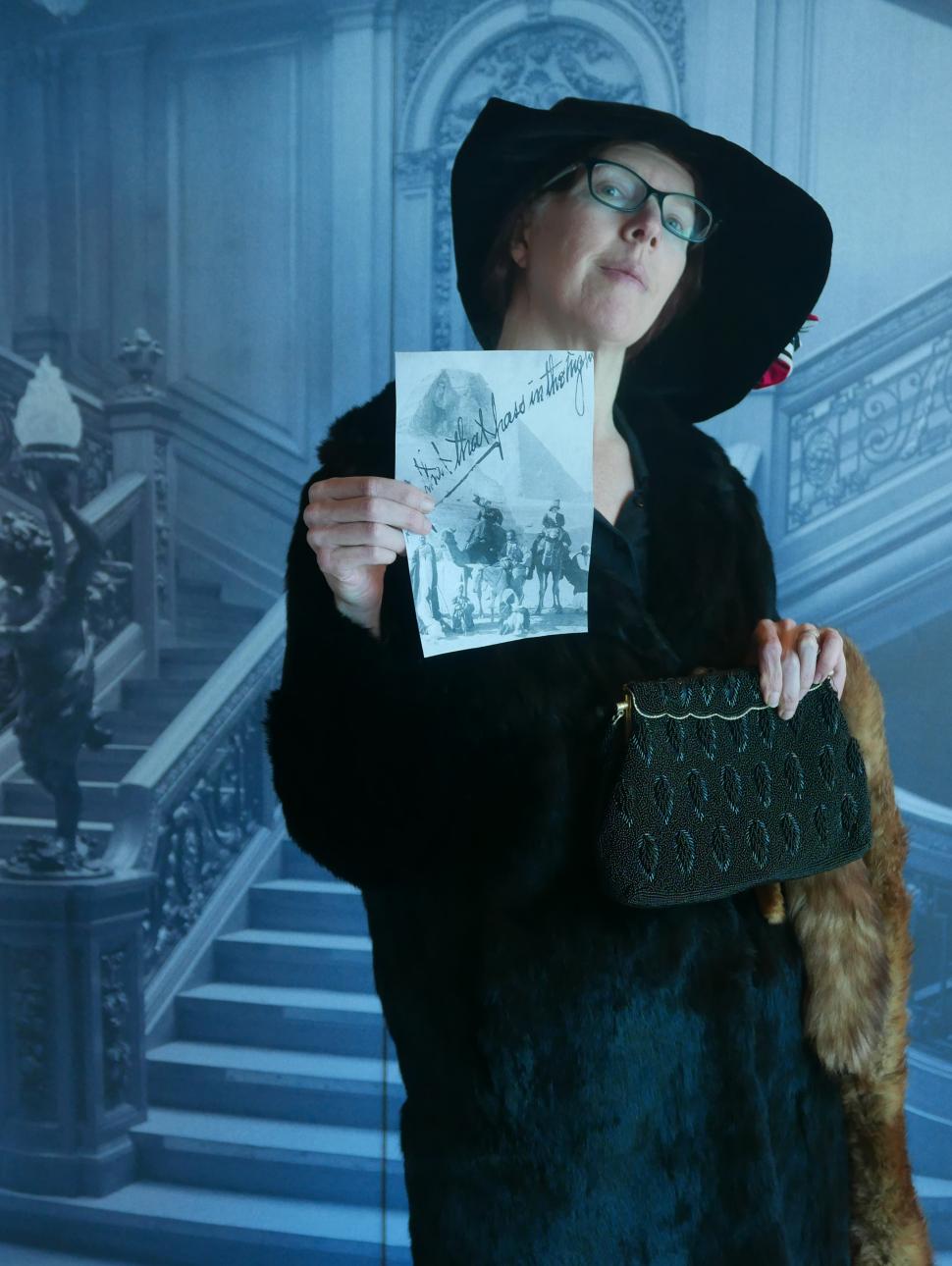 A woman dressed in early 20th century clothing stands in front of an ornate staircase, holding a photograph