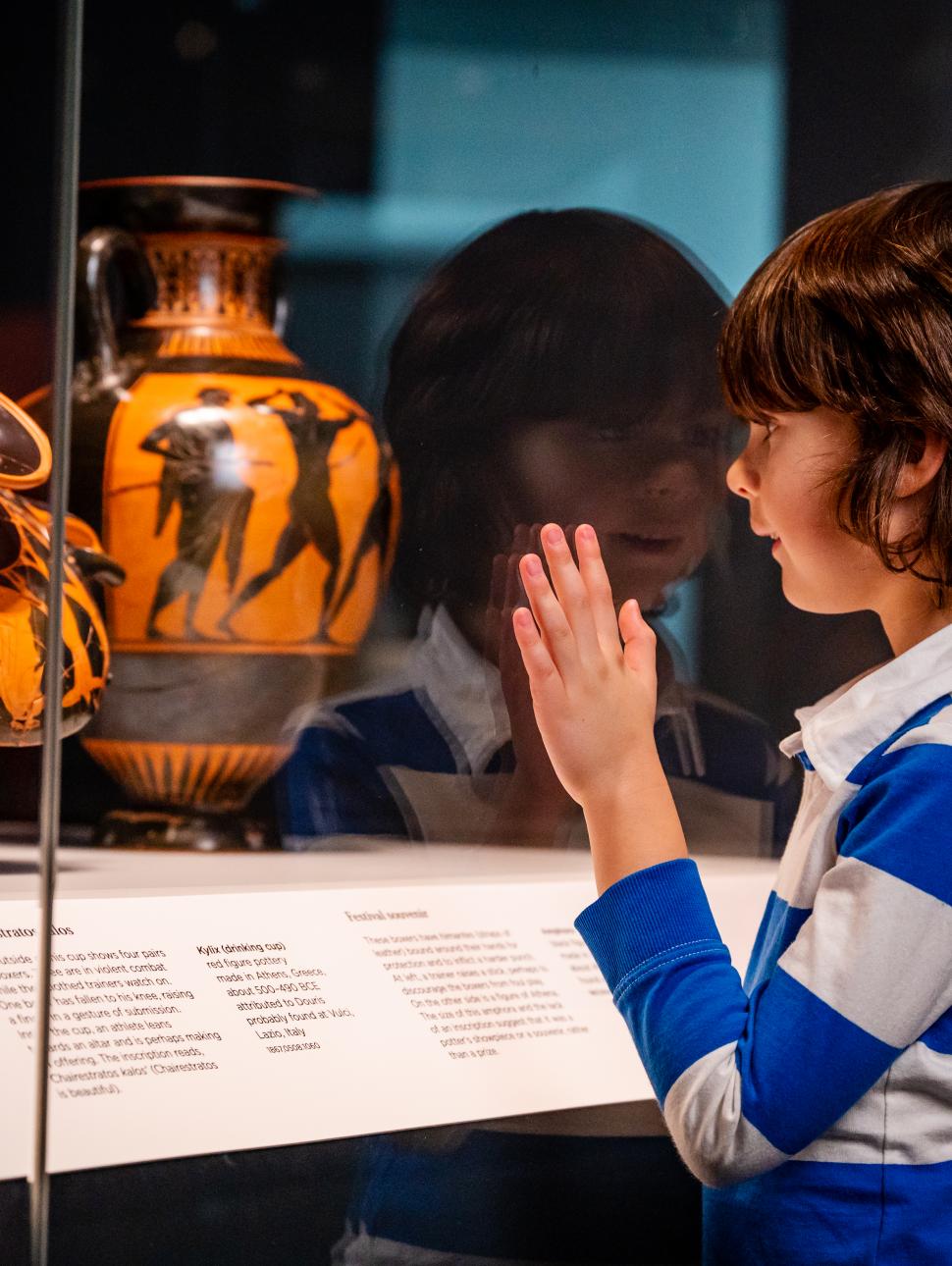 Child in a striped blue and white looking at an Ancient Greek ceramic piece displayed in a glass cabinet. 