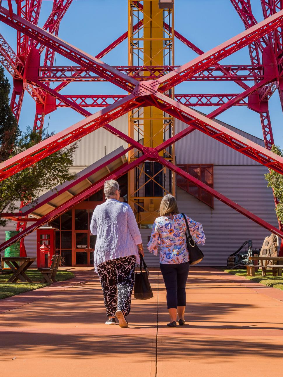 Two women walk away from the camera towards a bright red mining headframe