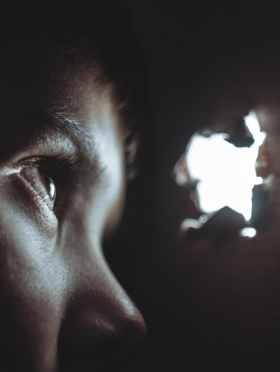 A close up view of a child in the dark peering through a small brightly lit hole in a wooden wall